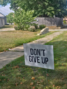 A white yard sign with black letters that read, "Don't Give Up." 