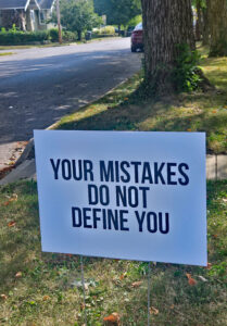 A white yard sign with black letters that read "Your mistakes do not define you."